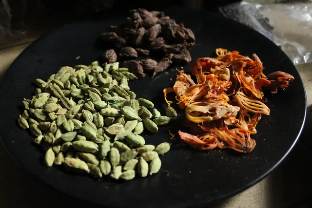 Black cardamom at the back of a plate with green cardamom and orange mace.