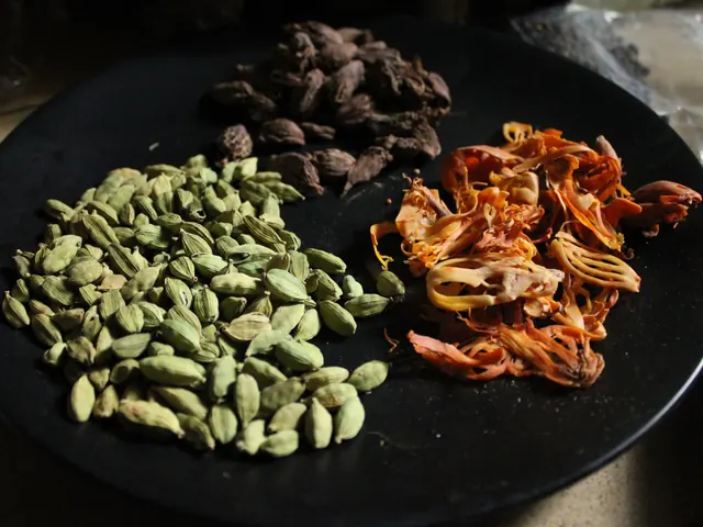 Black cardamom at the back of a plate with green cardamom and orange mace.